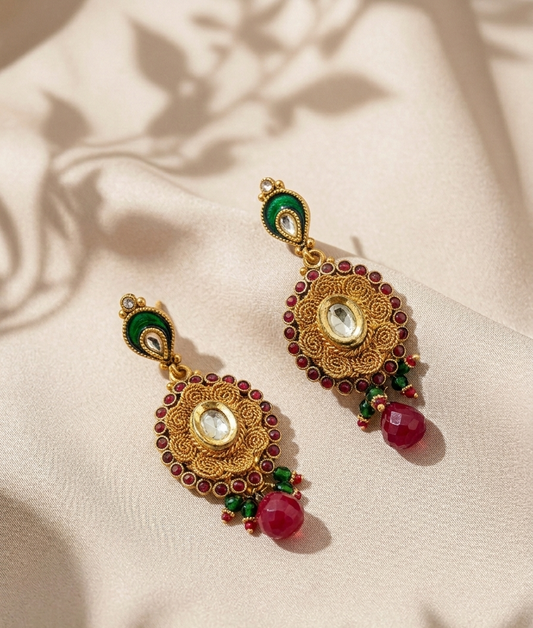 A pair of ornate gold-toned Indian-style earrings decorated with green enamel, red stones, and a clear central stone, hanging on a cream satin fabric with soft leaf shadows.