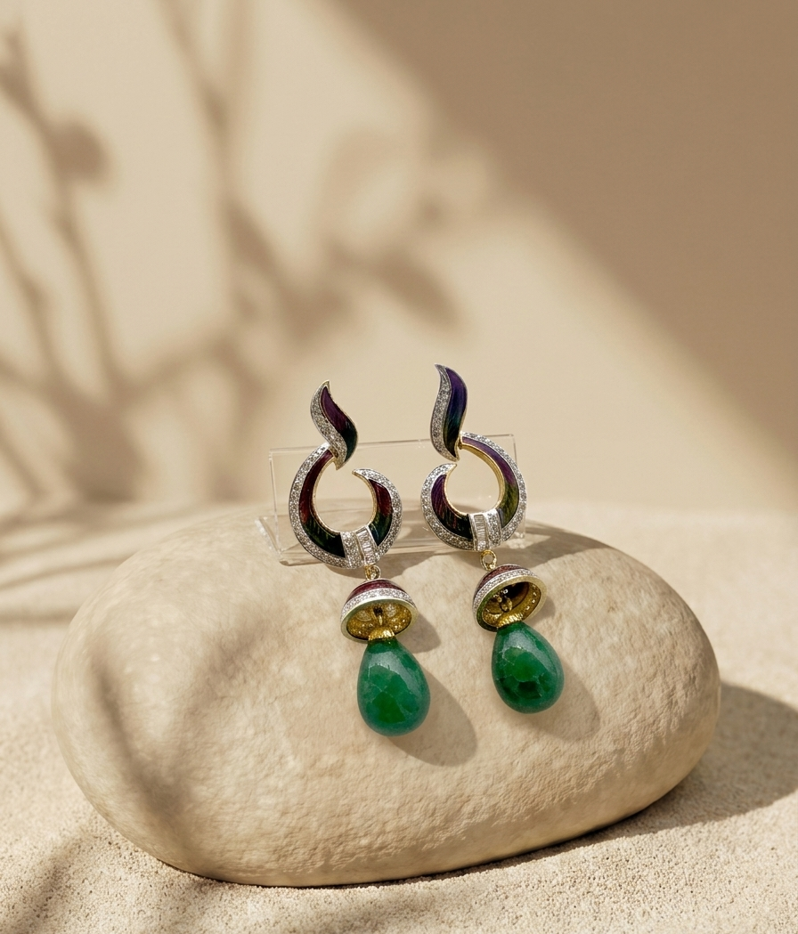 A pair of ornate dangle earrings displayed on a smooth beige stone. The earrings feature curved silver-toned hoops with dark purple and green enamel detailing, small clear stones, and a gold-toned circular accent above a teardrop-shaped green stone drop.