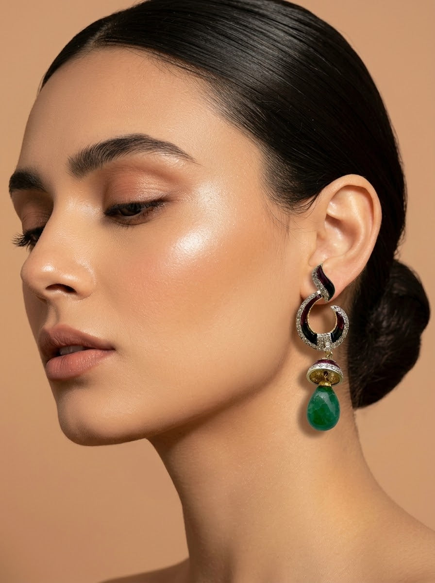 A close-up of a woman wearing a dramatic statement dangle earring. The earring features a black and silver crescent-shaped top set with small clear stones, a gold-toned accent, and a dangling green teardrop gemstone.