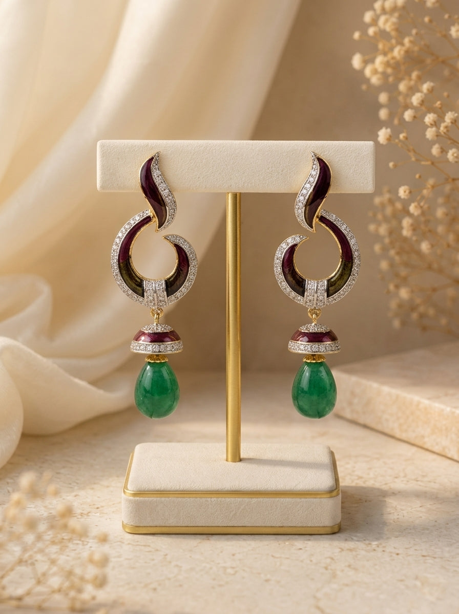 A pair of ornate drop earrings displayed on a cream jewelry stand. The earrings feature curved silver-toned hoops set with clear stones and deep red enamel, with a dangling green teardrop gemstone at the bottom.