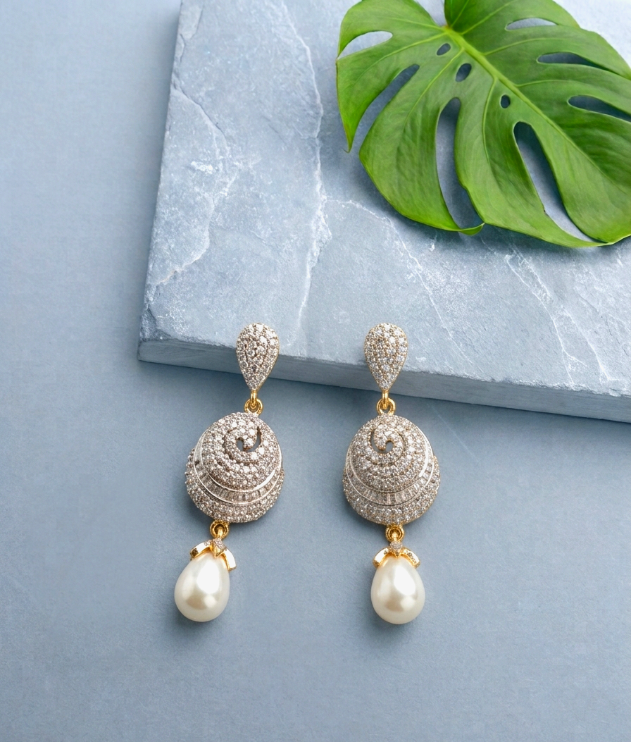 A pair of gold-plated drop earrings adorned with pavé-set clear stones and dangling white pearls, displayed on a light blue-gray surface next to a green monstera leaf and a gray stone slab.