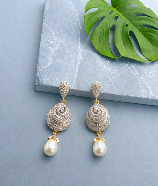 A pair of gold-plated drop earrings adorned with pavé-set clear stones and dangling white pearls, displayed on a light blue-gray surface next to a green monstera leaf and a gray stone slab.