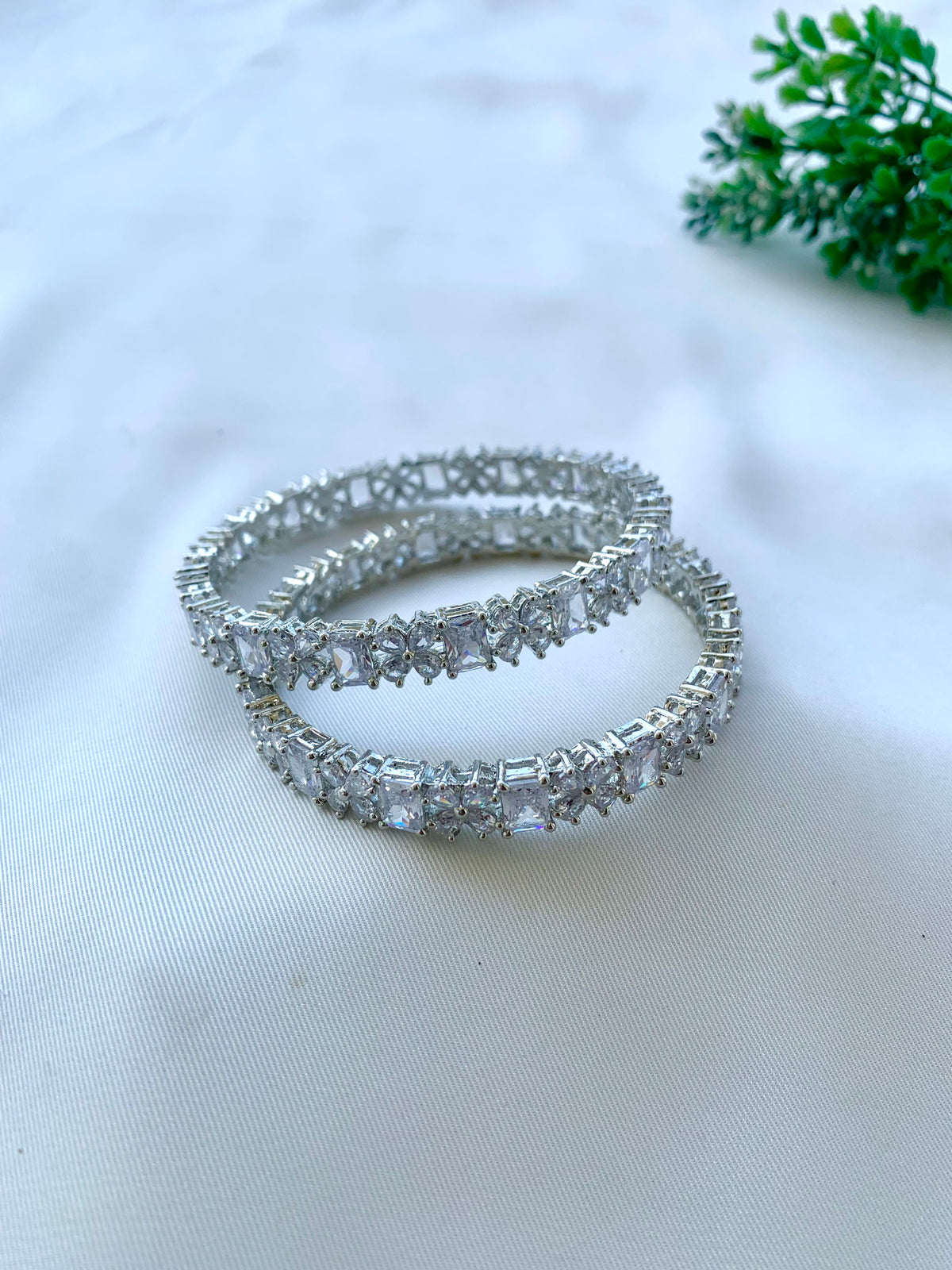Two silver-toned bangle bracelets adorned with clear rectangular gemstones, arranged on a white fabric surface with a small green plant in the top right corner.