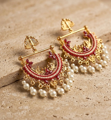 Ornate gold earrings with red stones and pearl-like beads on a textured beige surface