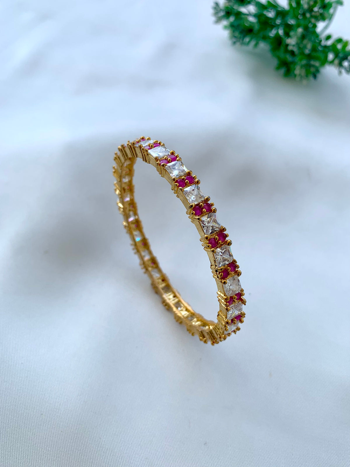 A gold eternity-style ring with rectangular clear stones and rows of small pink gemstones, photographed on a soft white fabric background with a small green plant in the corner.