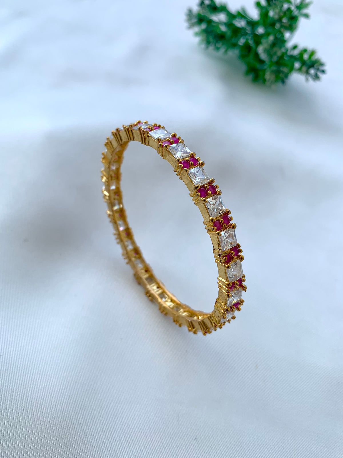 A gold eternity ring featuring baguette-cut clear stones and small red gemstones, displayed on a soft white fabric background with a small green plant in the background.