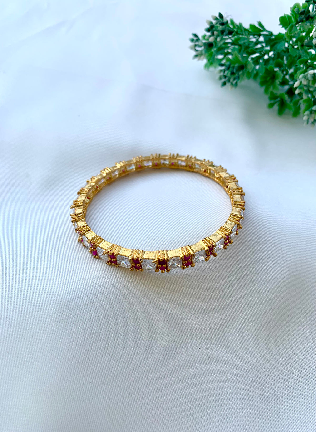 A gold-plated bangle bracelet adorned with clear rectangular crystals and small red stones, displayed on a white fabric background with a green plant in the corner.