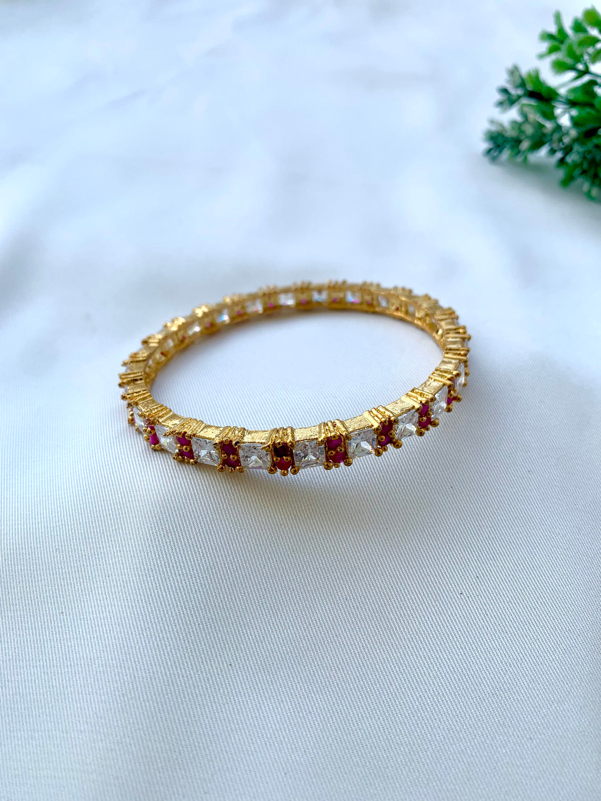 A gold-plated bangle bracelet featuring a row of square clear crystals and small red stones, displayed on a white textured fabric background with a small green plant in the corner.