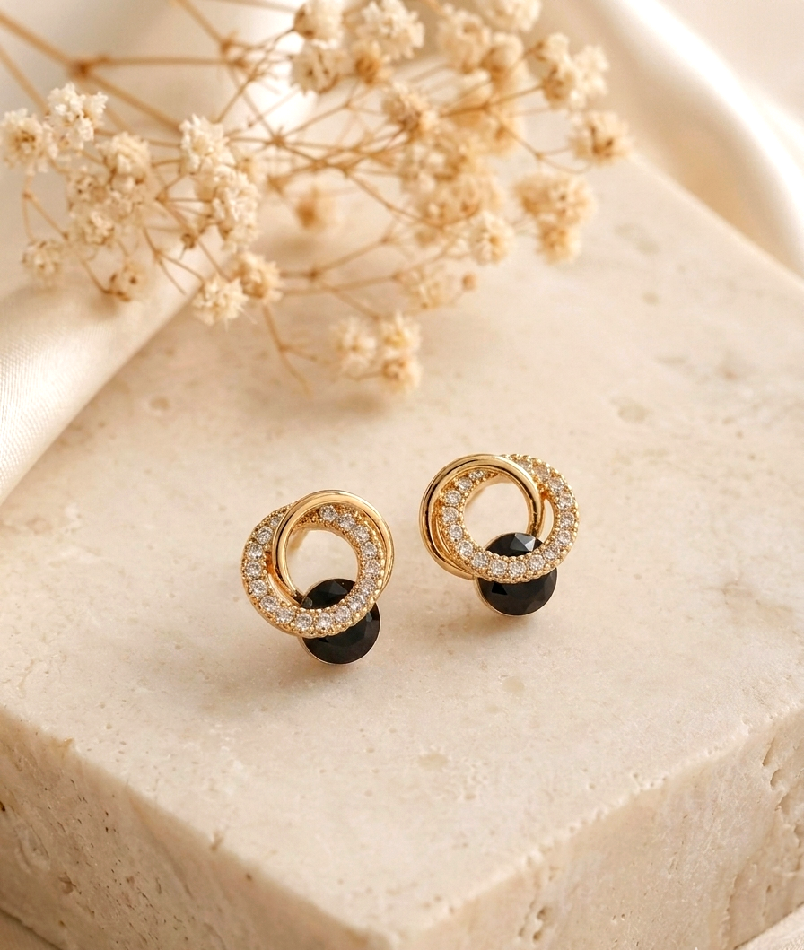A pair of gold-toned double-ring stud earrings with clear crystal detailing and a small black bead drop, displayed on a beige stone block with dried flowers in the background.