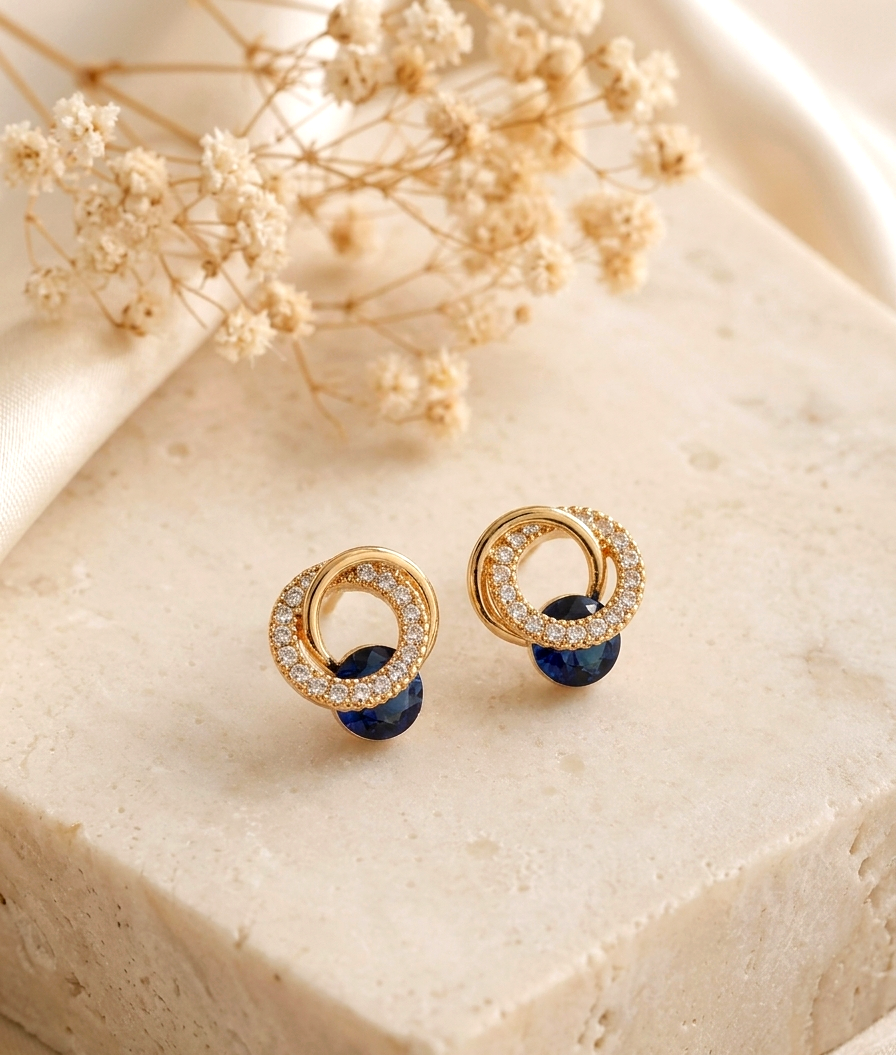 A pair of gold-toned stud earrings with double interlocking rings, one adorned with small clear crystals and the other with dark blue stones, displayed on a textured beige stone block with dried flowers in the background.