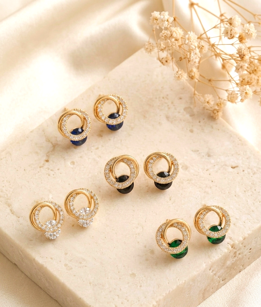 Six pairs of gold hoop stud earrings arranged on a cream stone slab with dried flowers. The earrings feature either clear pavé-set stones, dark navy blue beads, or emerald green beads.