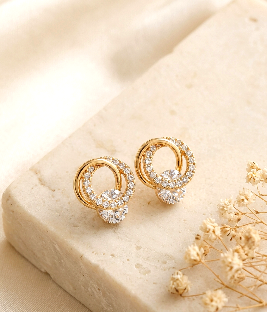 A pair of gold hoop earrings with clear crystal accents, displayed on a beige stone block with dried flowers.