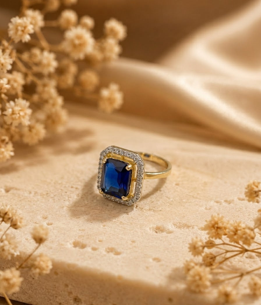 A gold ring with a rectangular blue gemstone surrounded by small clear stones, displayed on a beige stone surface with dried flowers and soft fabric in the background.
