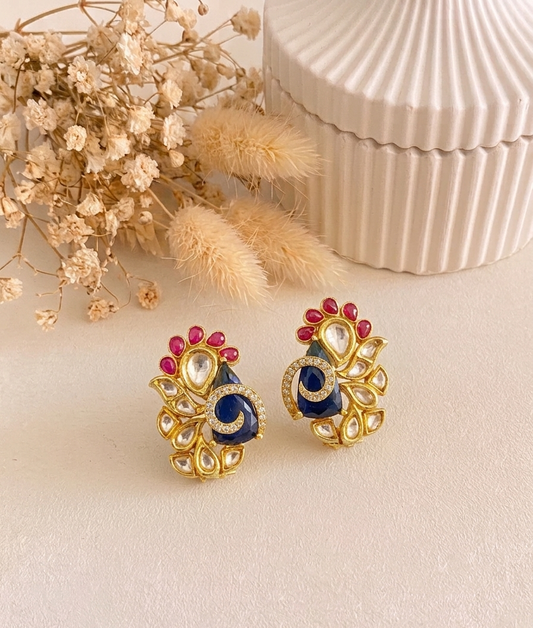 A pair of gold-toned Indian-style earrings with peacock-inspired designs, featuring deep blue teardrop centers, red gemstone accents, and clear stone details, displayed on a beige surface with dried flowers and a ribbed cream box in the background.
