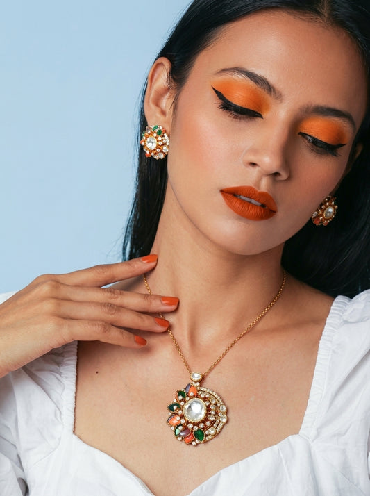 A close-up of a woman wearing a gold necklace with a large, ornate circular pendant featuring white pearls and green and orange-red stones, matching drop earrings, and orange makeup against a light blue background.