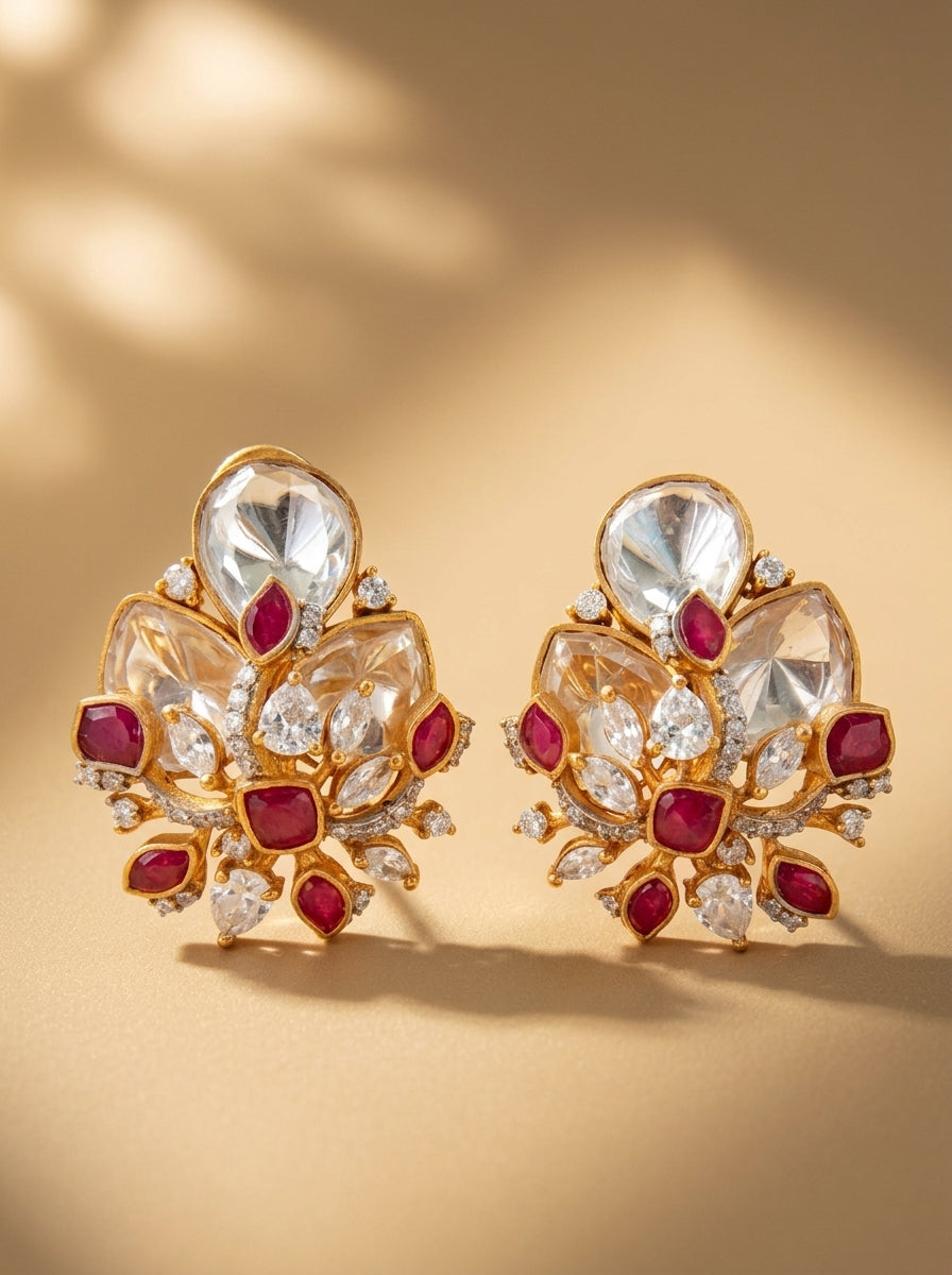 A pair of gold-toned statement earrings featuring large clear stones and red gemstone accents, displayed on a warm beige background with soft shadows.