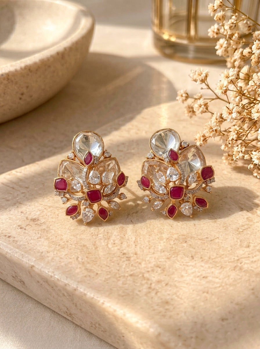 A pair of gold-toned statement earrings with clear teardrop-shaped crystals and small red gemstone accents, displayed on a beige stone surface with dried flowers and a bowl in the background.