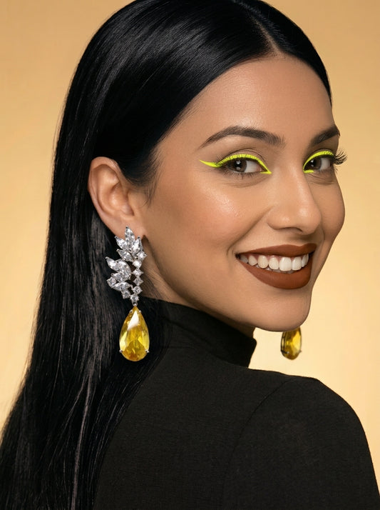 A close-up portrait of a woman with long black hair wearing a black turtleneck and large statement earrings. The earrings feature clear gemstone clusters with teardrop-shaped yellow-gold drops.