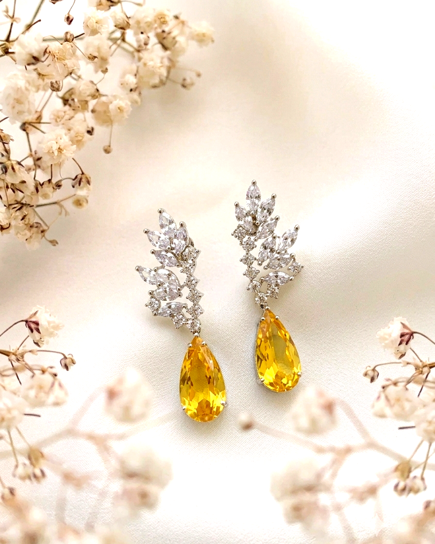 A pair of silver-toned drop earrings with clear crystal leaf-shaped tops and yellow teardrop gemstones, styled on a soft cream fabric with dried white flowers.