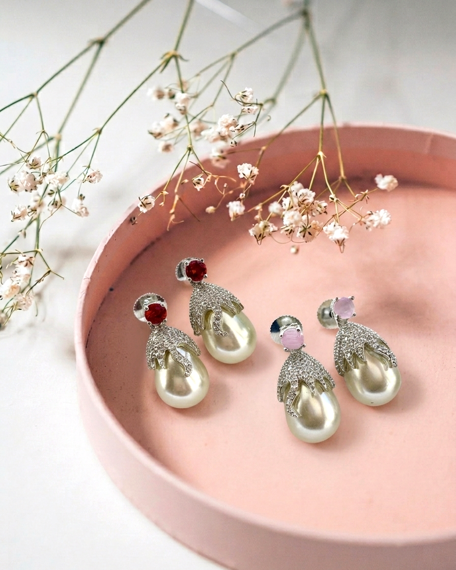 Four pairs of pearl and crystal earrings arranged in a pink round tray with delicate white flowers on a light marble surface. The earrings feature large white pearls, silver crystal-encrusted leaf-like settings, and either red or light pink gemstones at the top.