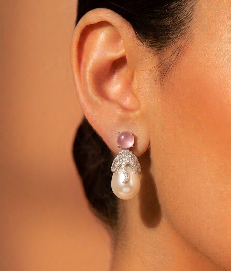 A close-up of a model's ear wearing a dangling earring with a pink cabochon stone, a white pearl drop, and a silver pavé setting.