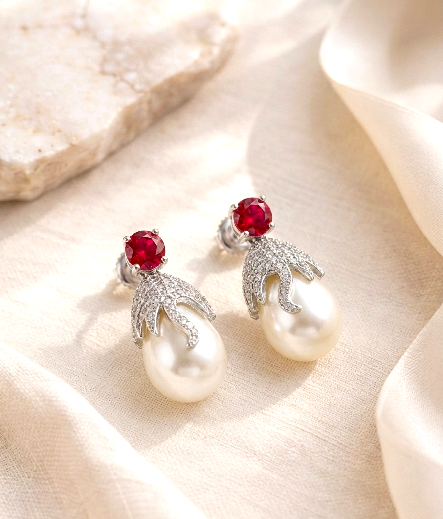 A pair of silver-toned drop earrings with round red gemstones and large white pearl drops, styled on draped cream fabric with a stone prop.