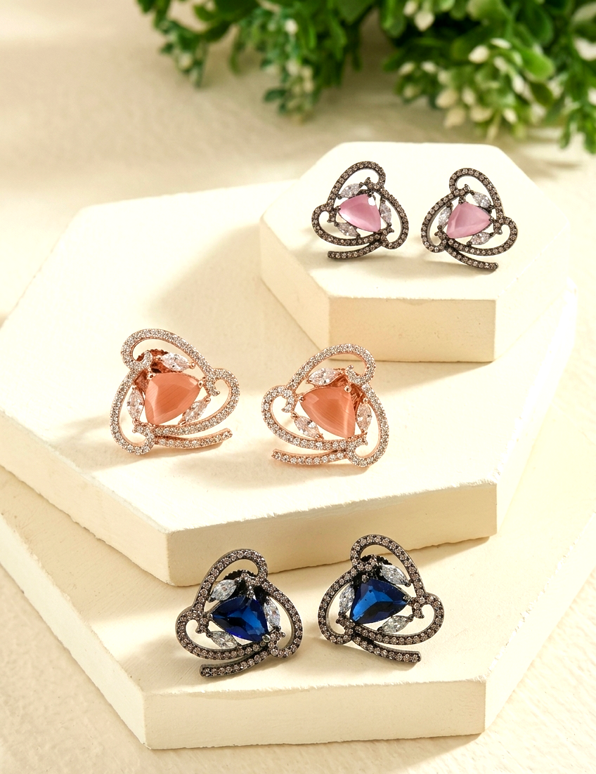 Three pairs of heart-shaped stud earrings displayed on cream geometric blocks. The earrings feature heart-shaped stones in pink, peach, and deep blue, surrounded by curved silver-toned and rose-gold pavé settings with small clear stones.