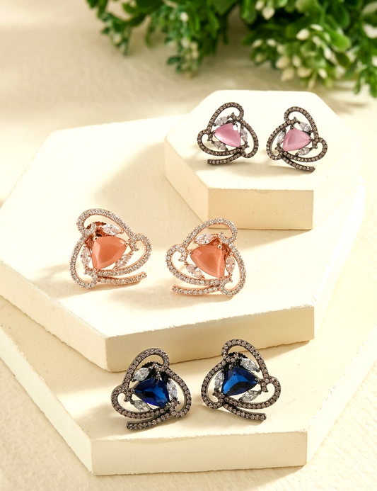 Three pairs of heart-shaped stud earrings displayed on cream geometric blocks. The earrings feature heart-shaped stones in pink, peach, and deep blue, surrounded by curved silver-toned and rose-gold pavé settings with small clear stones.