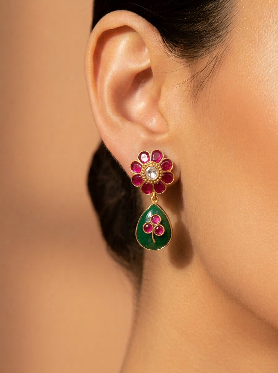 A close-up of a model's ear wearing a gold-toned traditional Indian floral dangle earring with pink enamel petals, a central clear stone, and a green teardrop-shaped drop with pink floral detailing.