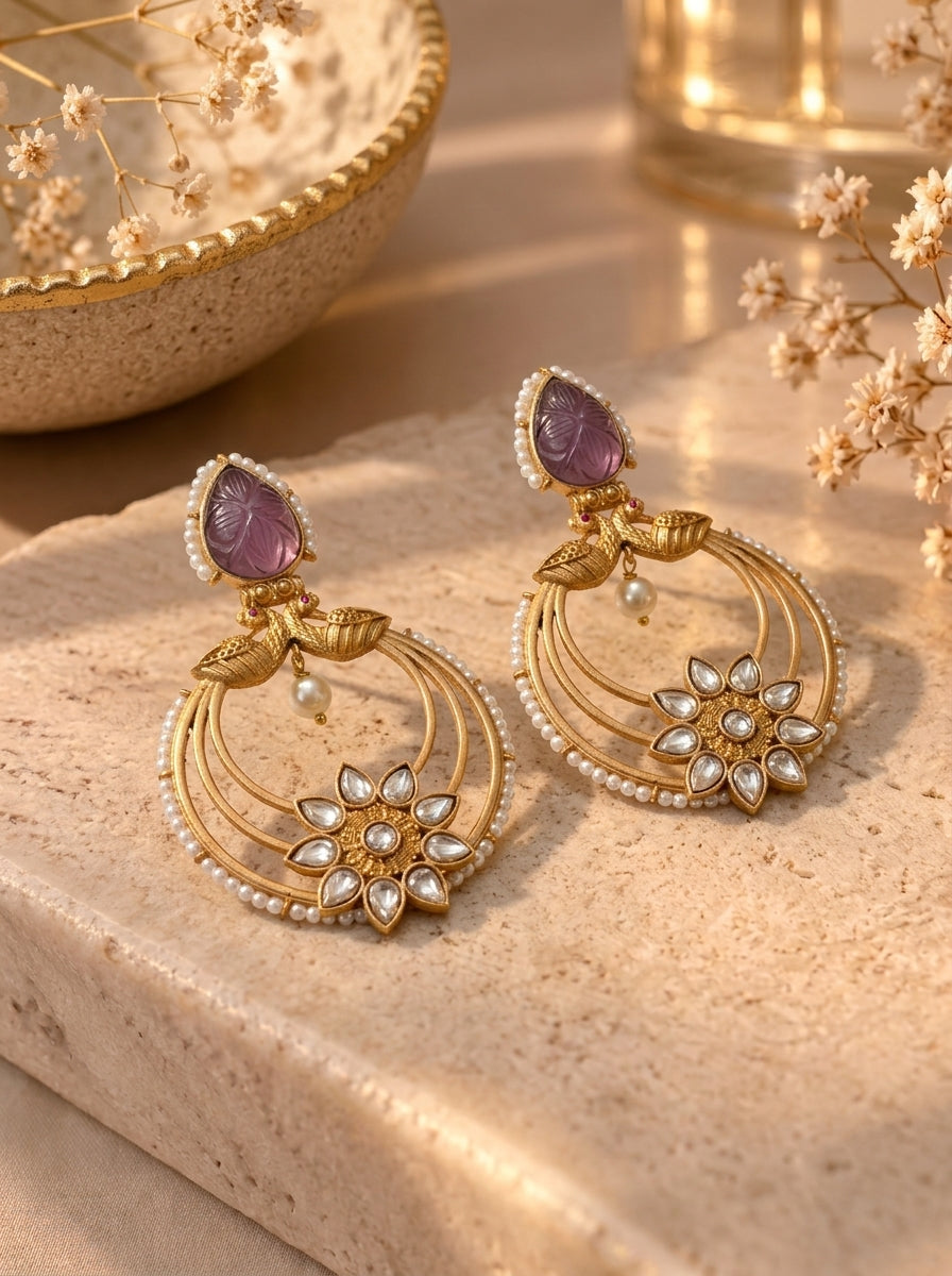 A pair of gold-toned Indian-style dangle earrings with purple amethyst stones, white pearl-like beads, and a floral motif, displayed on a beige stone slab with dried flowers.