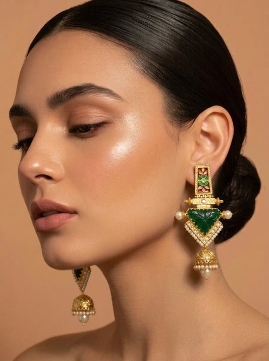 A close-up of a woman wearing a large gold-toned statement earring with a prominent green heart-shaped stone and smaller green and white accents, against a warm beige background.