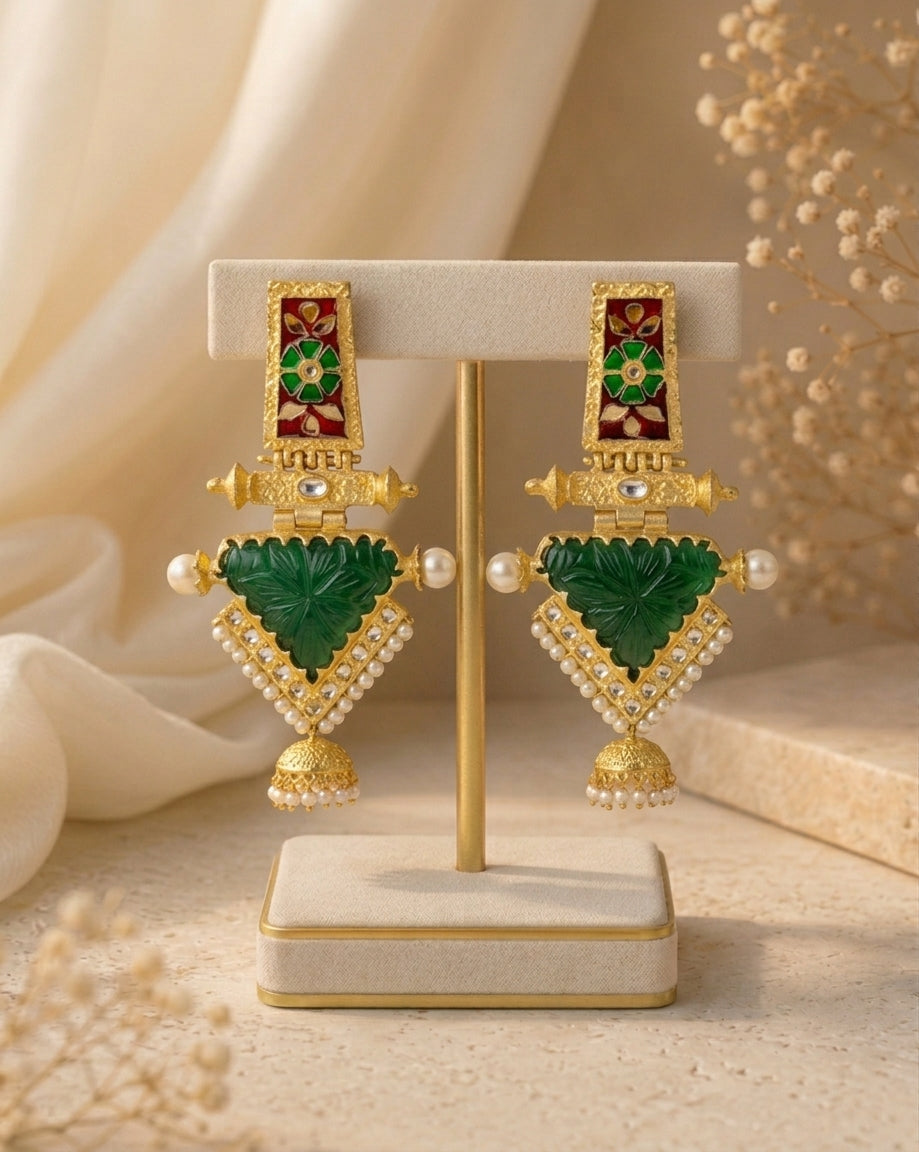 A pair of ornate gold-toned traditional earrings displayed on a cream jewelry stand. The earrings feature green stone inlays, small white pearl-like beads, and red and green enamel details at the top, set against a softly styled beige background with draped fabric and dried floral decor.