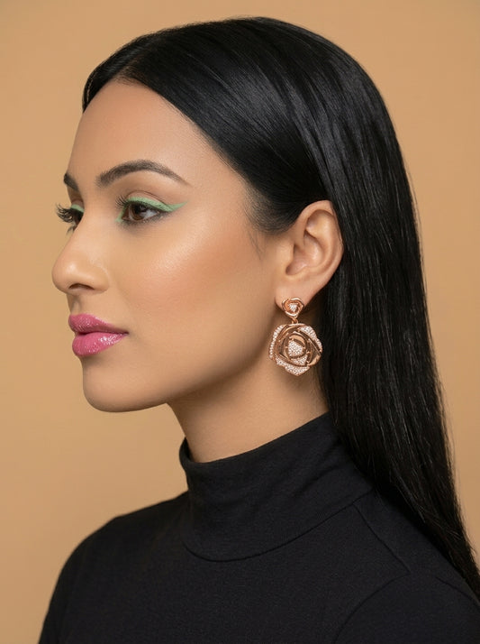 A woman in profile wearing a rose gold rose-shaped dangle earring with a white pearl center, set against a solid beige background.