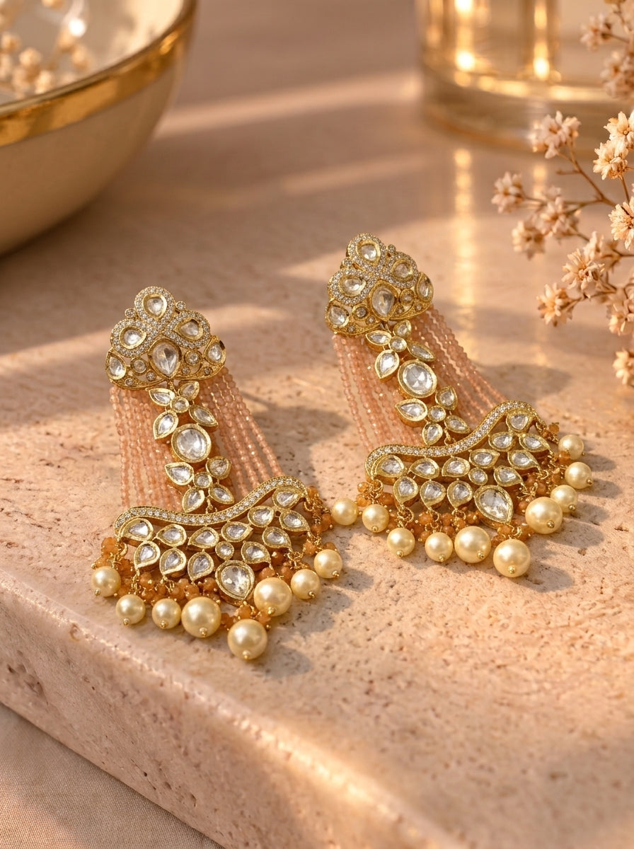 A pair of ornate gold-toned traditional Indian earrings with clear stones and pearl-like beads, displayed on a textured beige stone surface with warm sunlight and decorative dried flowers.