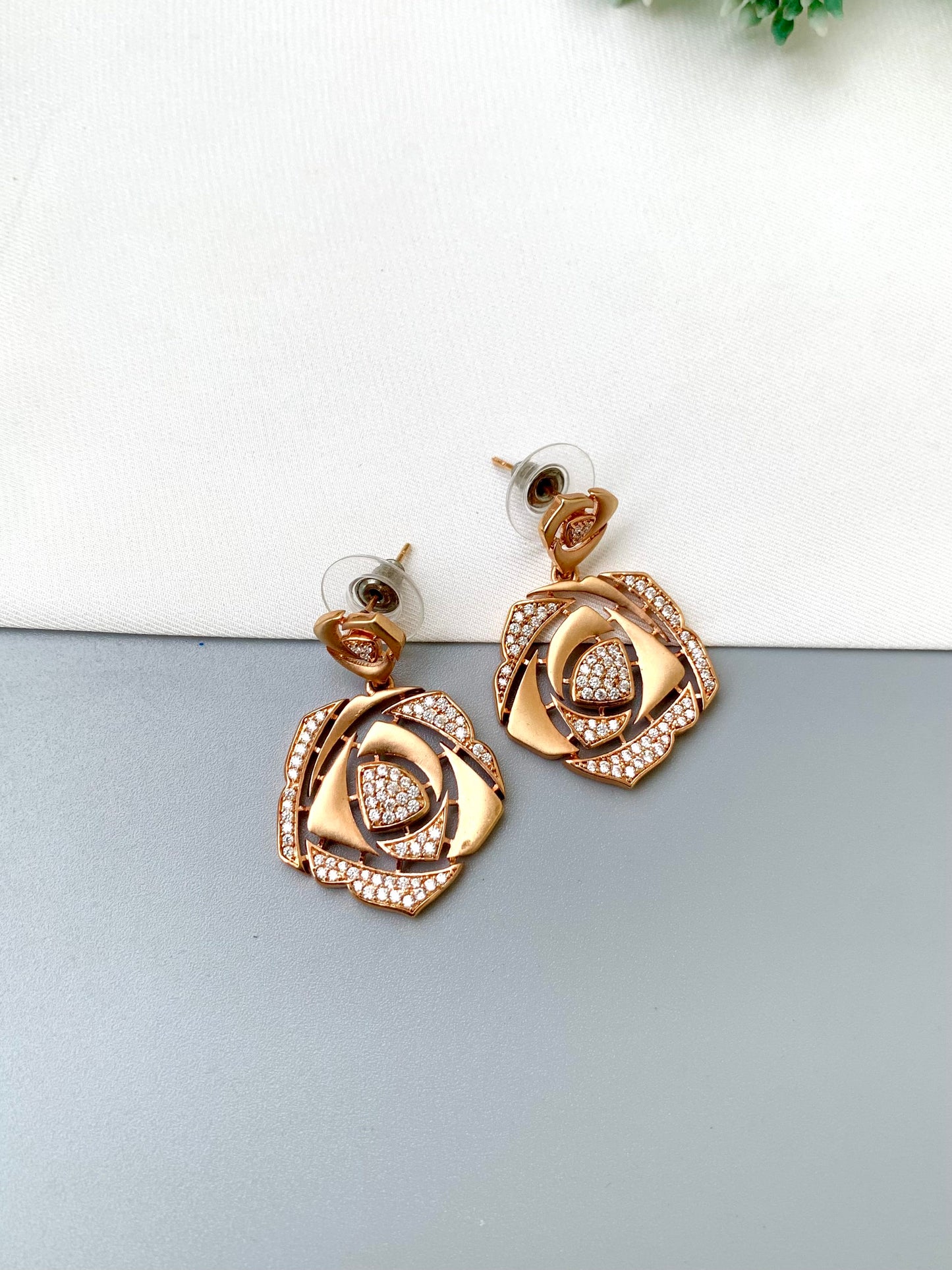 Rose gold rose-shaped drop earrings with pavé crystal detailing, displayed on a split white and gray background.