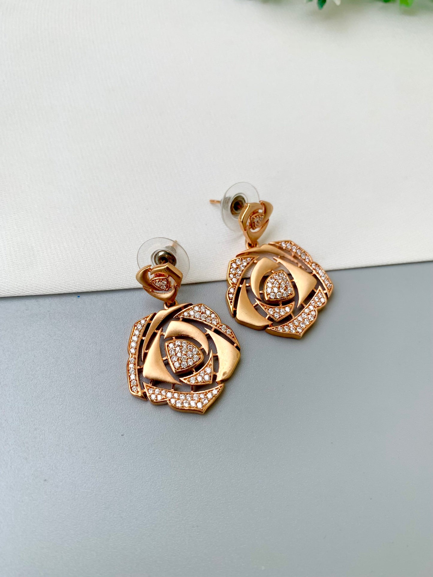 A pair of rose gold rose-shaped drop earrings with pavé crystal accents, displayed on a split white and gray surface.