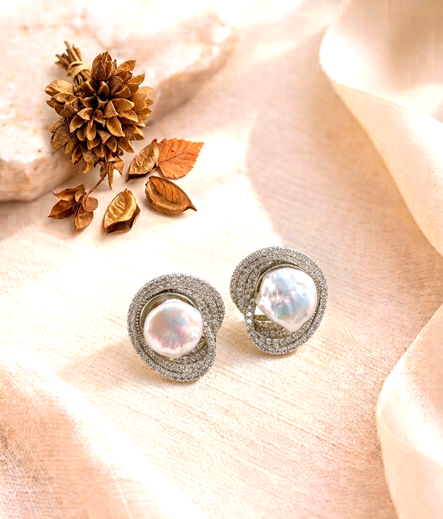 A pair of silver-toned stud earrings with large iridescent baroque pearls and a halo of small clear stones, styled on soft beige fabric with dried leaves and a stone.