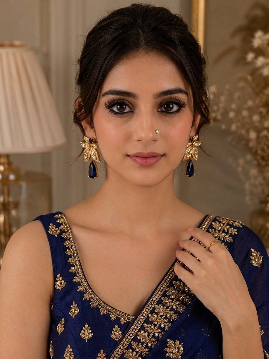 A woman wearing a navy blue embroidered traditional blouse with gold detailing, styled with matching gold and navy earrings, in an indoor setting with a lamp in the background.
