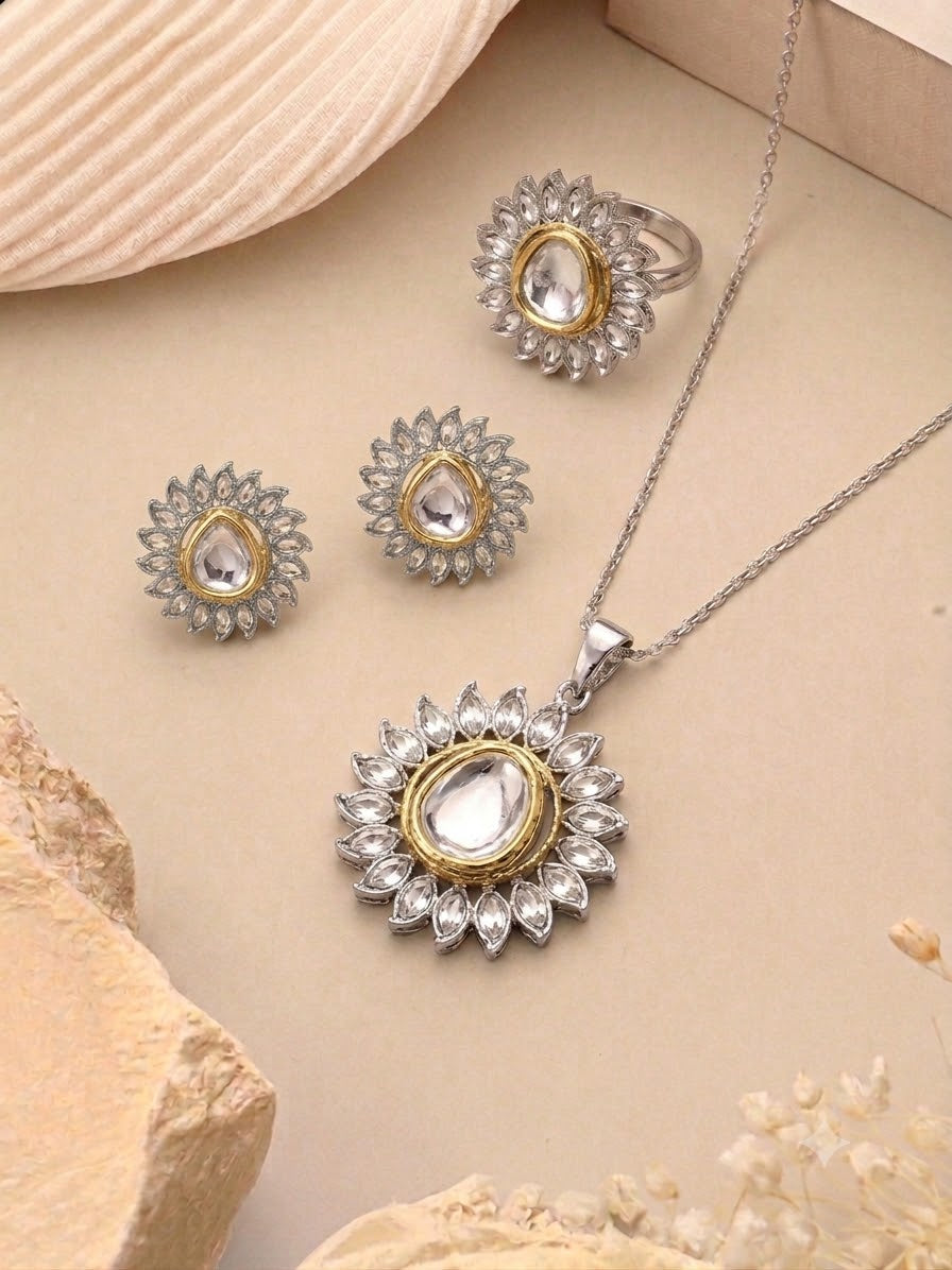 A silver and gold jewelry set featuring a pendant necklace, matching earrings, and a ring, arranged on a beige textured background with decorative seashell and stone props.