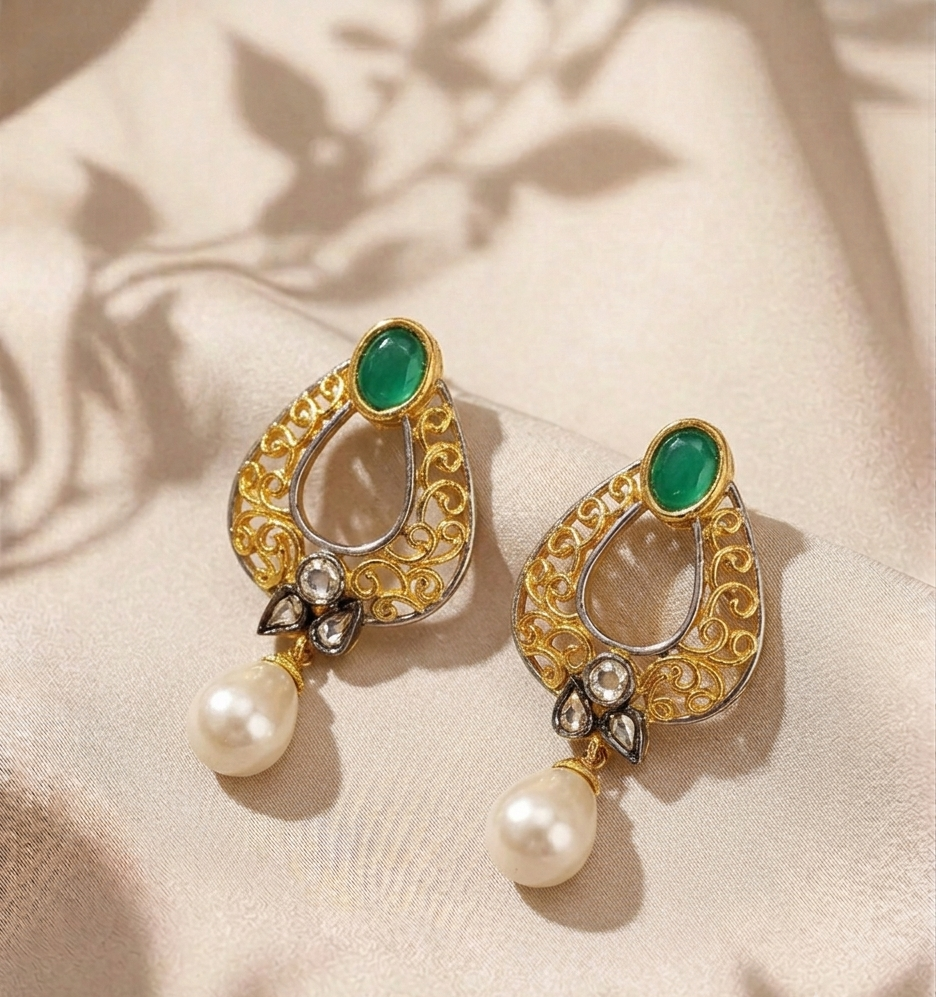 A pair of ornate gold filigree earrings with green oval stones and dangling white pearls, displayed on a beige satin fabric with soft leaf shadows.