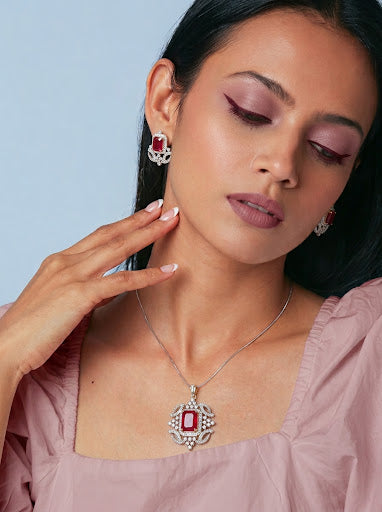 A woman wearing a silver pendant necklace and matching stud earrings with rectangular red gemstones, styled with a dusty pink top against a light blue background.