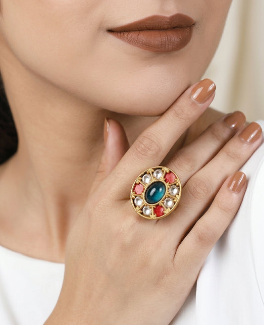 A close-up lifestyle shot of a model wearing a large gold ring with a central blue-green stone and surrounding red and white stones.