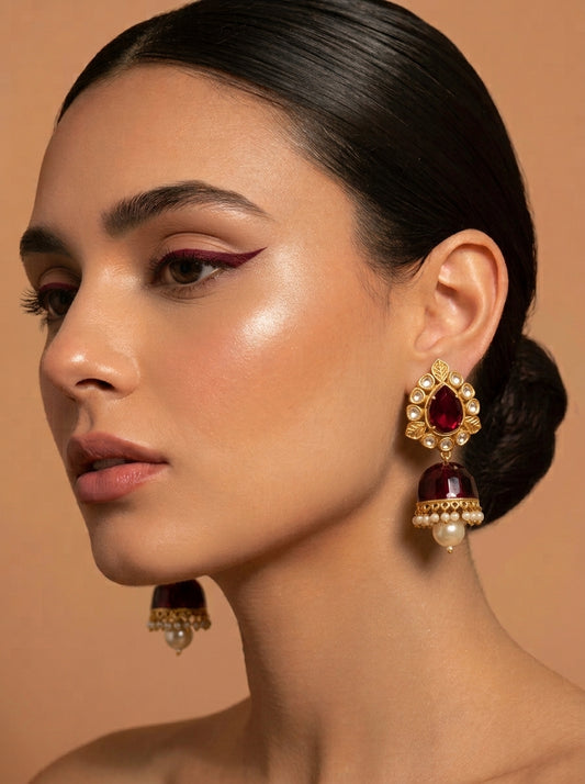 A close-up of a woman with dark hair pulled back, wearing a pair of ornate gold-toned earrings with deep red teardrop stones and small pearl-like accents, against a warm beige background.
