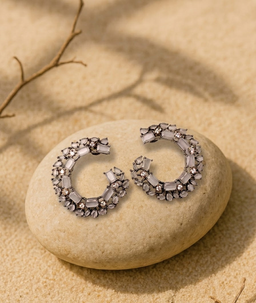 A pair of silver crescent-shaped hoop earrings adorned with clear faceted crystals, displayed on a smooth beige stone with a sandy background and soft leaf shadows.