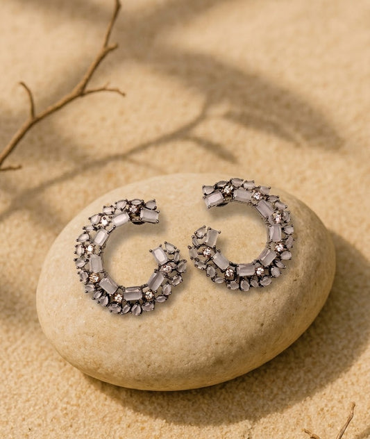 A pair of silver crescent-shaped hoop earrings adorned with clear faceted crystals, displayed on a smooth beige stone with a sandy background and soft leaf shadows.