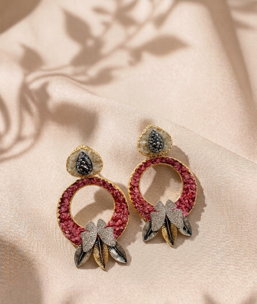 A pair of gold-plated drop earrings with textured pink circular hoops, crystal-encrusted teardrop studs, and dangling crystal and gold leaf motifs, displayed on a beige satin fabric with soft shadows.