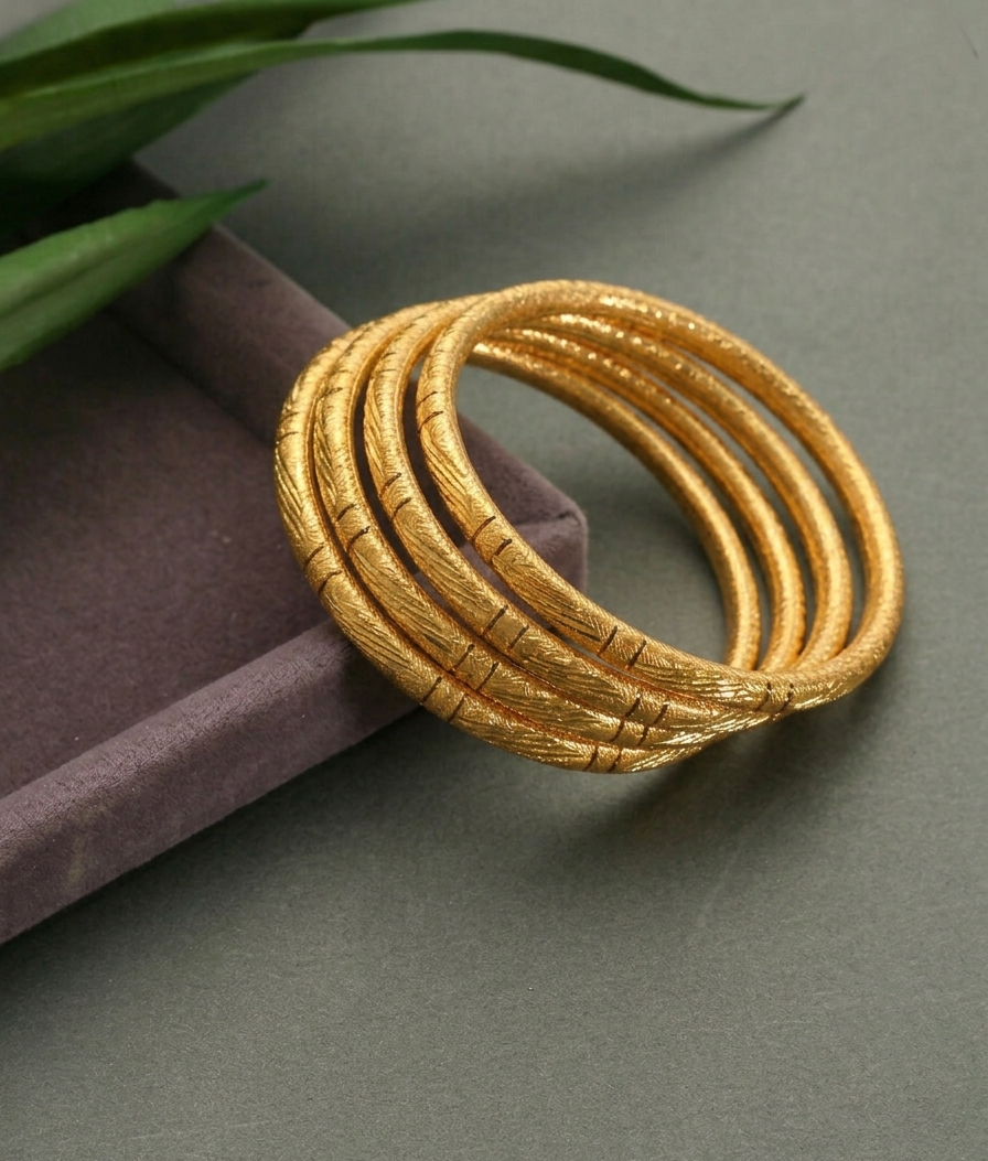 A stack of five textured gold bangles resting on a dark purple velvet box, with green leaves in the top-left corner against a dark gray background.