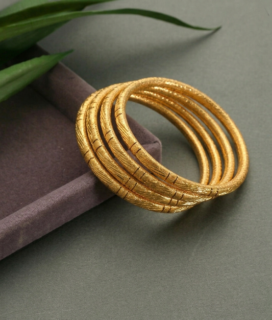 A stack of five textured gold bangles resting on a dark purple velvet box, with green leaves in the top-left corner against a dark gray background.