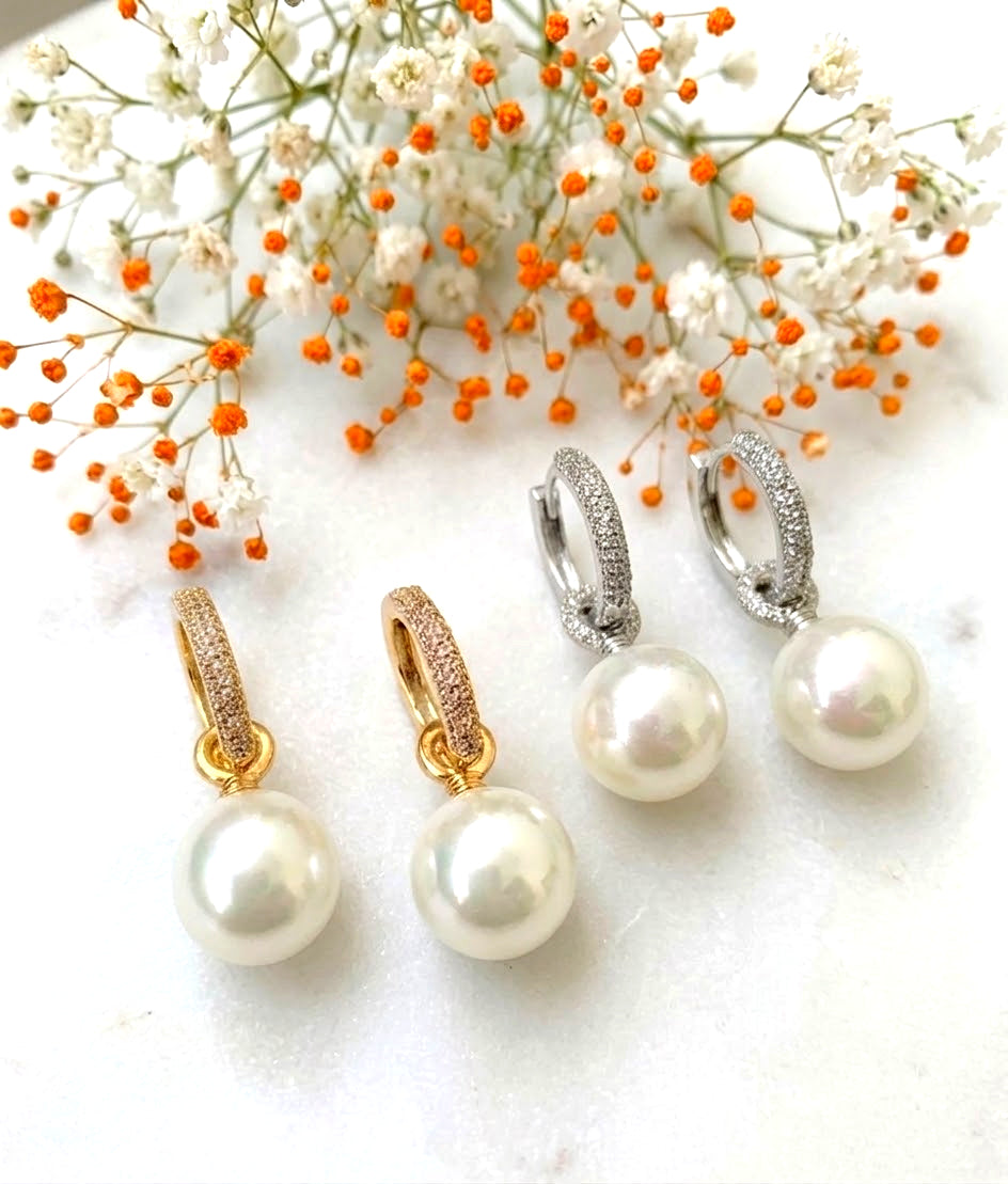 Four pearl drop earrings with huggie hoops, shown in gold, rose gold, silver, and white gold settings, styled with baby's breath flowers on a white marble surface.