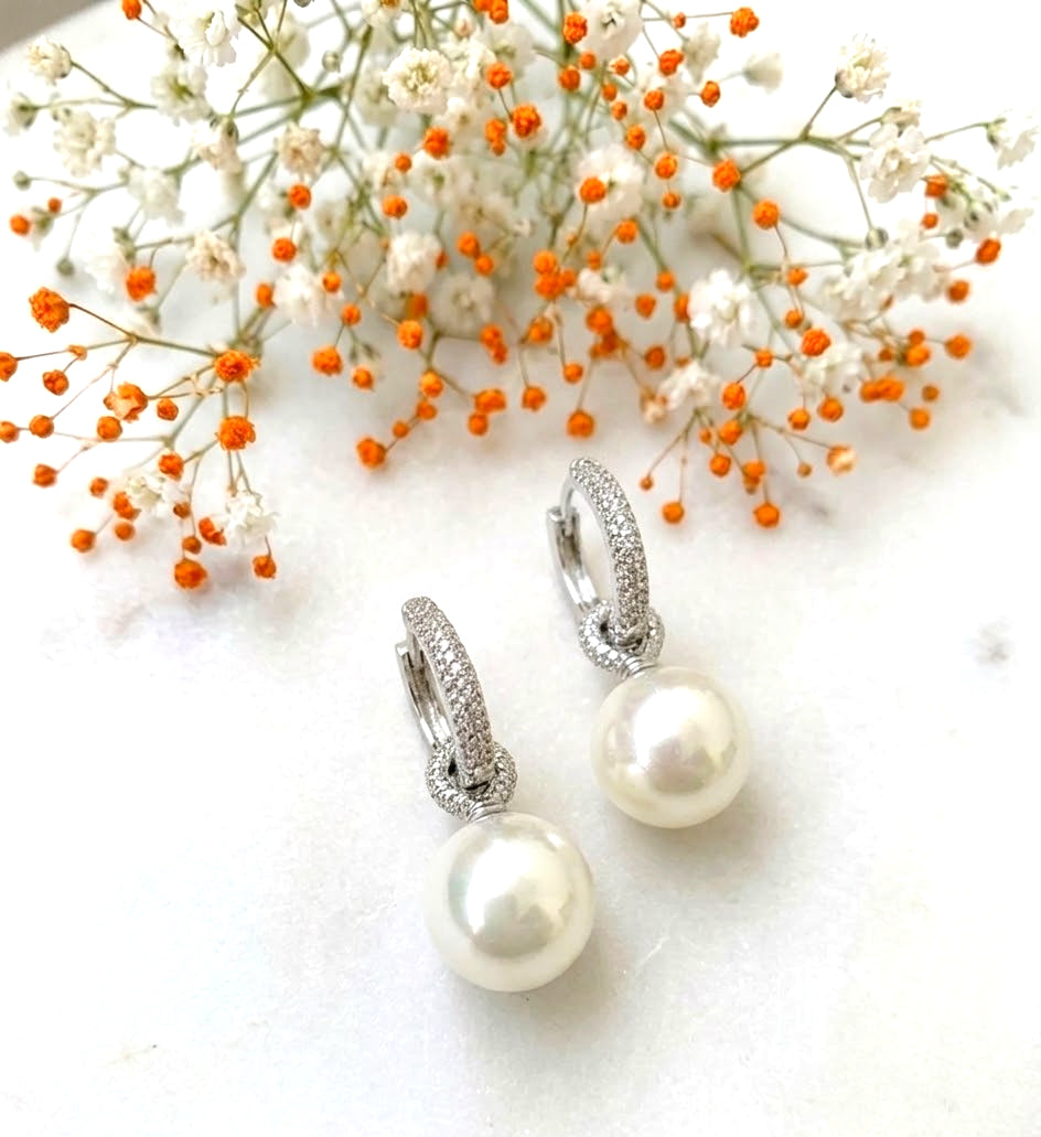 A pair of silver hoop earrings with a pavé-set knot design and a dangling white pearl, styled on a white marble surface with baby's breath flowers.